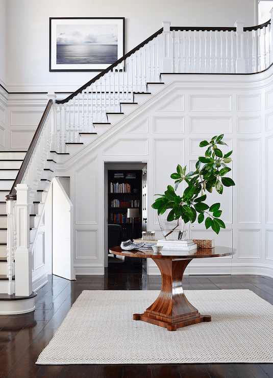 Stunning foyer features full wall wainscoting hiding door leading to secret room situated behind round entry table atop West Elm Jute Chenille Herringbone Rug layered over glossy wide plank floors.