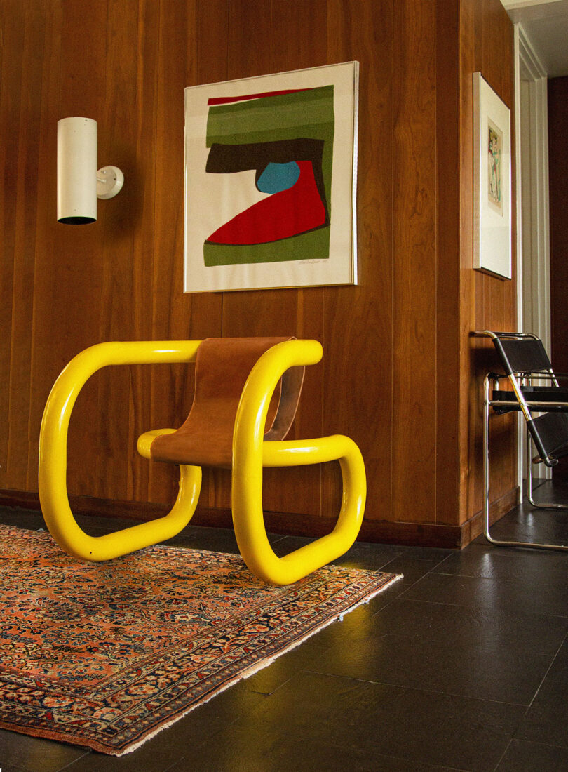 A contemporary room featuring a yellow tubular chair on a patterned rug, with modern art on the wood-paneled walls.