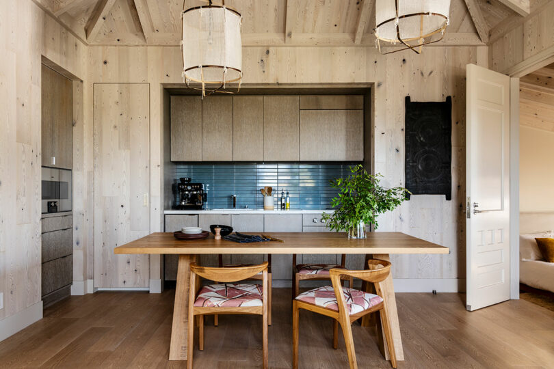 Modern kitchen interior designed by SkB Architects featuring a wooden dining table, chairs, light wood paneling, and elegant overhead lighting.