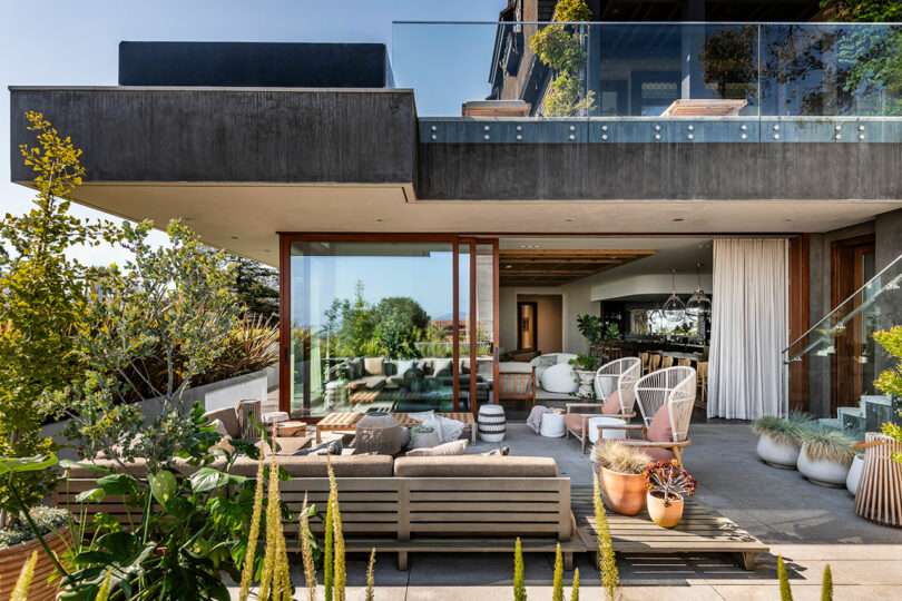 Modern house designed by SkB Architects with expansive glass doors open to a patio furnished with a sofa and chairs, overlooking a garden and distant hills.