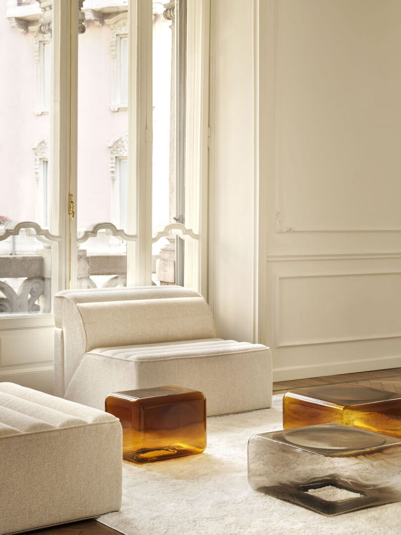 Elegant living room with a white sofa and three transparent glass side tables in shades of amber and grey on a patterned wood floor.