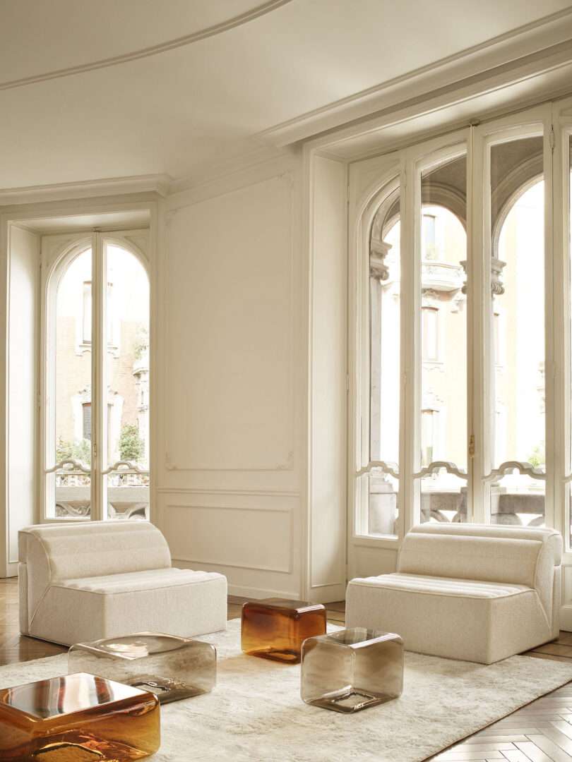 Elegant living room with a white sofa and four transparent glass side tables in shades of amber and grey on a patterned wood floor.