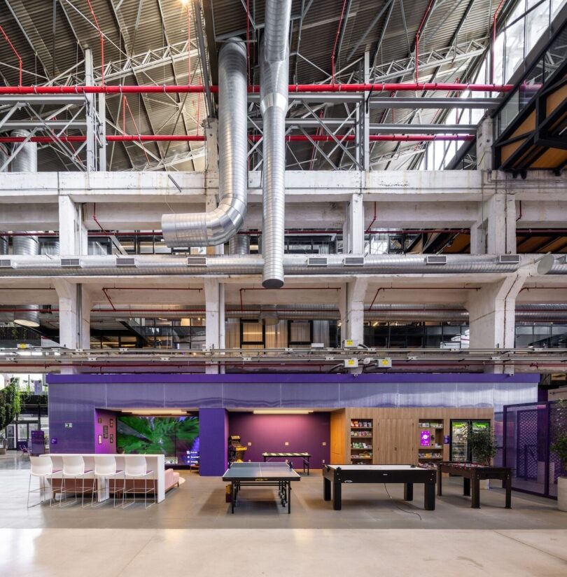 Spacious modern office with exposed ceiling pipes, colorful cubicles, and a lounge area with benches and a large screen display