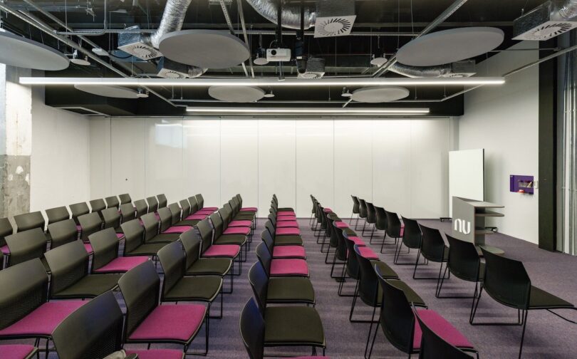 Modern conference room with rows of black chairs, whiteboard, and a projector on a gray ceiling with exposed HVAC system