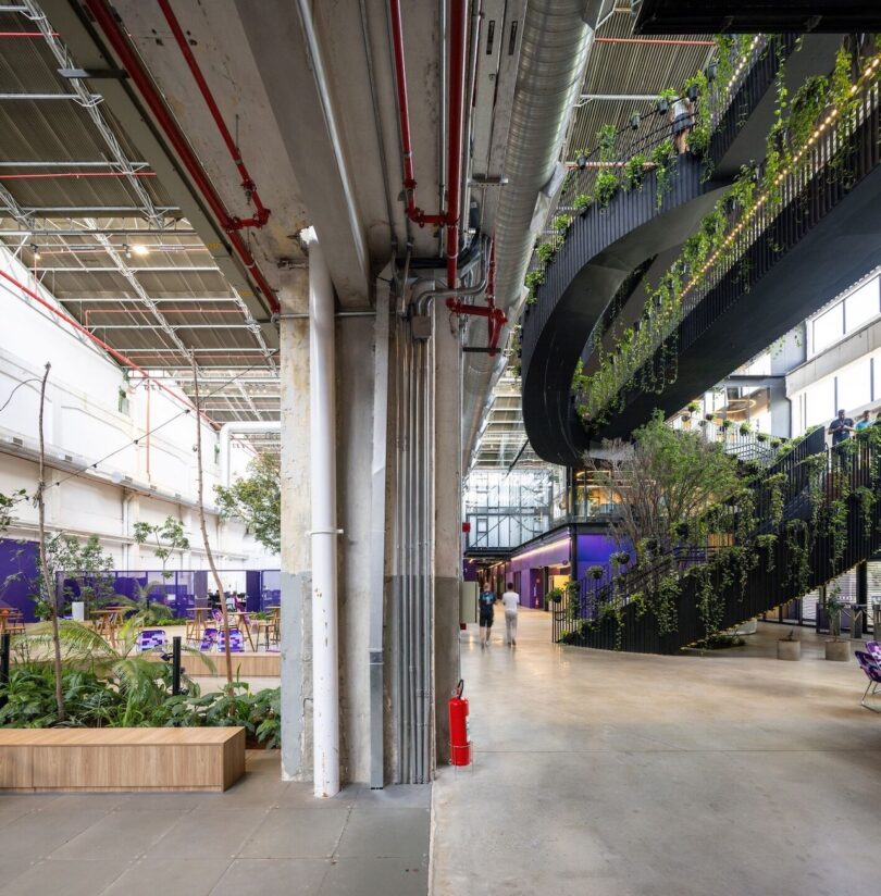 Interior view of a modern office building with exposed beams and pipes, featuring a large column, staircases, and hanging green plants