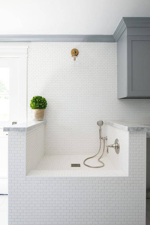 White mini brick tiles in a mudroom dog shower completed with a marble deck, a chrome shower head and gold wall mount single sconce.