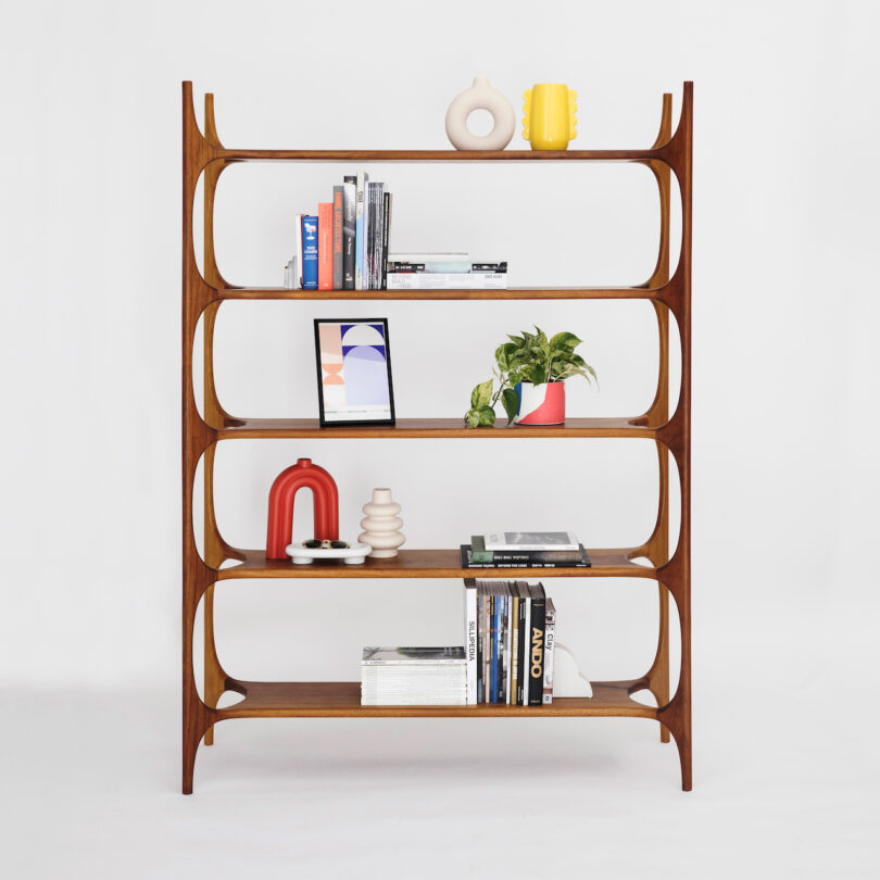 A wooden bookshelf with a curved design, holding various books, houseplants, and decorative items against a white background