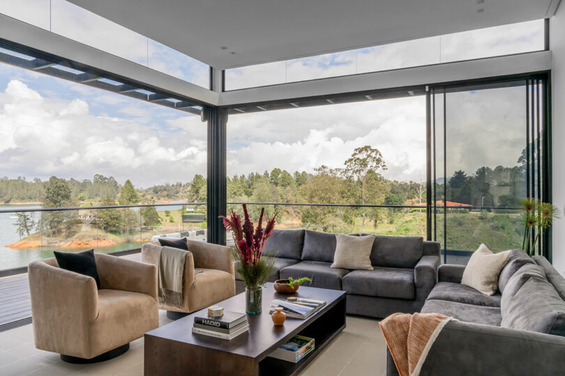 Modern living room with large windows overlooking a lake, featuring a gray sectional sofa, two beige armchairs, and a wooden coffee table.