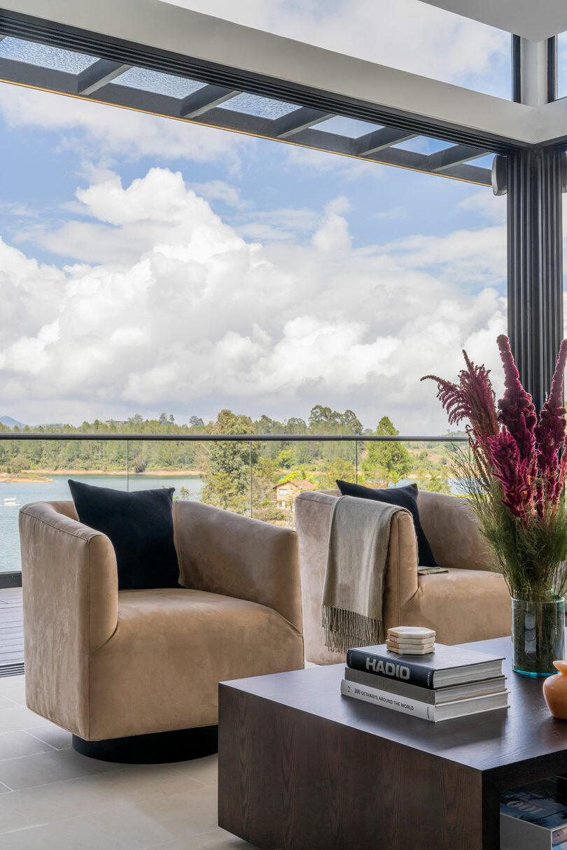 Modern living room interior with large windows showcasing a scenic lake view, featuring a plush beige armchair and a cozy throw blanket near an elegant coffee table.