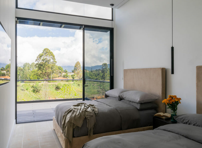 Modern bedroom with a large window overlooking a green landscape, featuring a neatly made bed with grey bedding and a small bedside table with books and flowers.