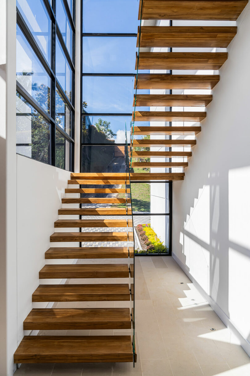 A modern staircase with wooden steps and glass railings in a bright room with large windows and sunlight casting shadows.