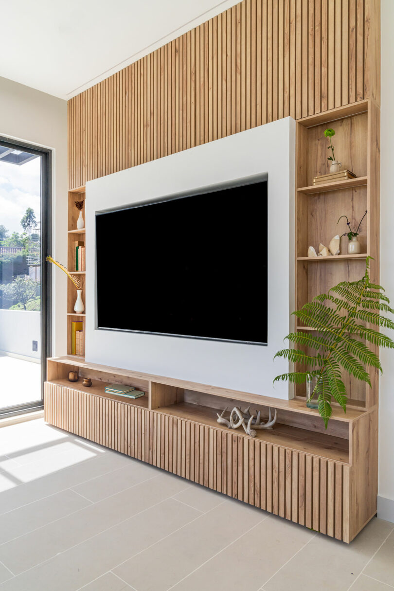 Modern living room with a large flat screen tv mounted on a wooden slatted wall with built-in shelving and a potted plant.