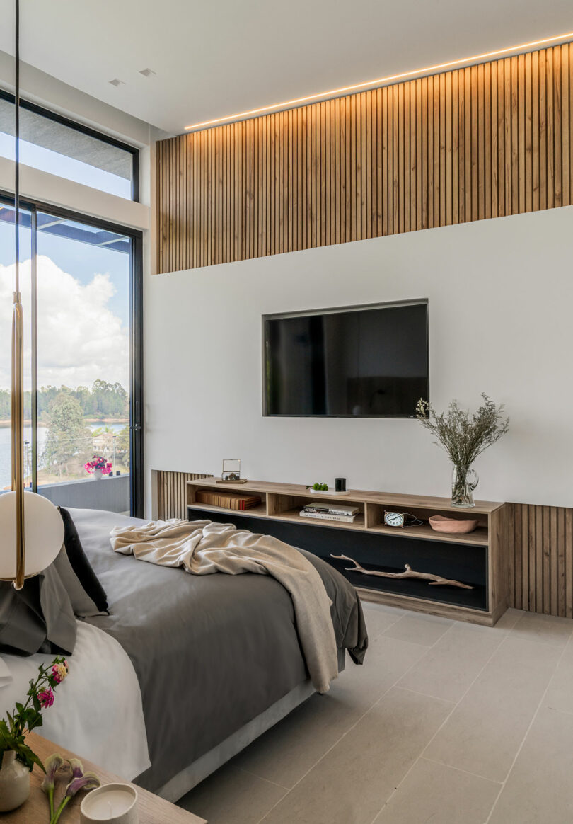 Modern bedroom with a large bed, wall-mounted tv, wooden slatted wall decor, floating shelves, and a large window.