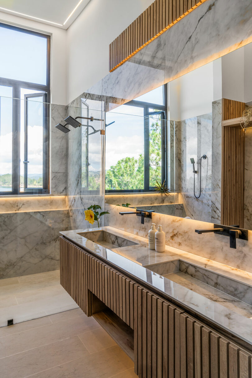 Modern bathroom interior with marble walls and wooden cabinets, featuring a double vanity, large mirrors, and a walk-in shower.