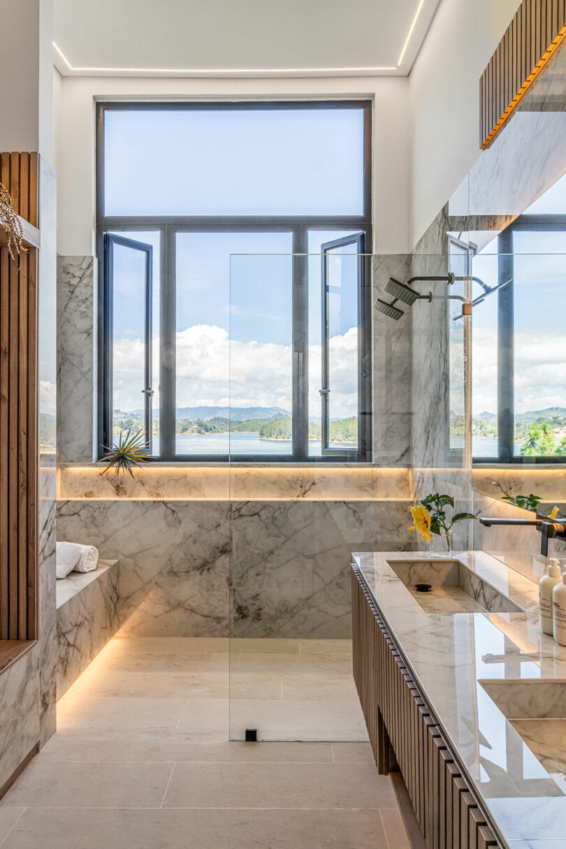 Luxurious marble bathroom with a glass shower, large windows overlooking a scenic view, and a double vanity.