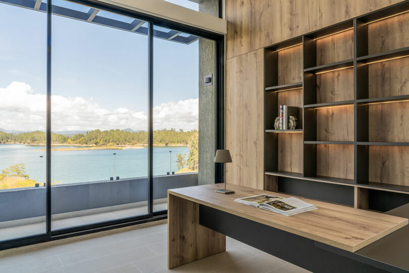 Modern office space with a large desk and built-in shelves, overlooking a scenic lake view through floor-to-ceiling windows.