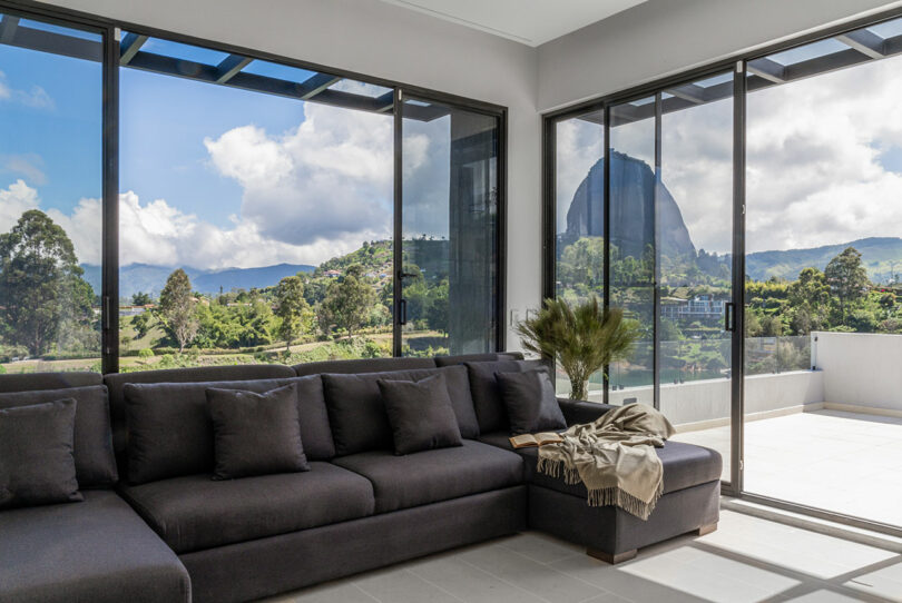 A modern living room with a large sectional sofa in front of floor-to-ceiling windows showcasing a panoramic view of a lush, mountainous landscape.