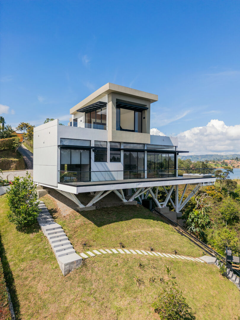 Modern house on a hillside featuring a large, angular design with glass facades, supported by steel beams, overlooking a lake under a clear sky.