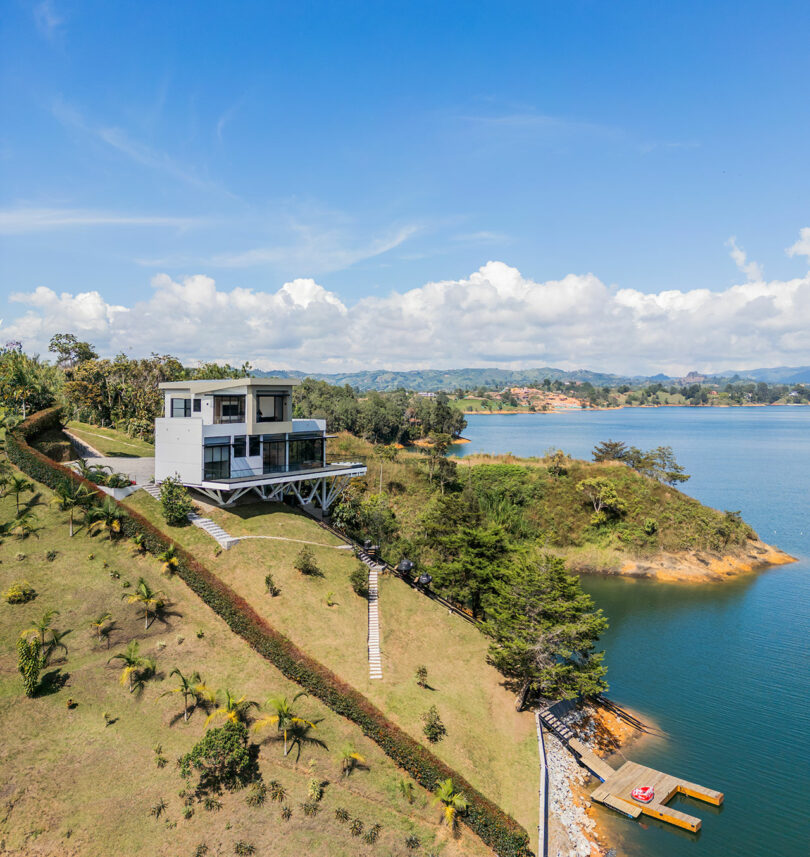 A modern two-story house on a landscaped hill overlooking a large lake, with a private dock extending into the water under a clear blue sky.