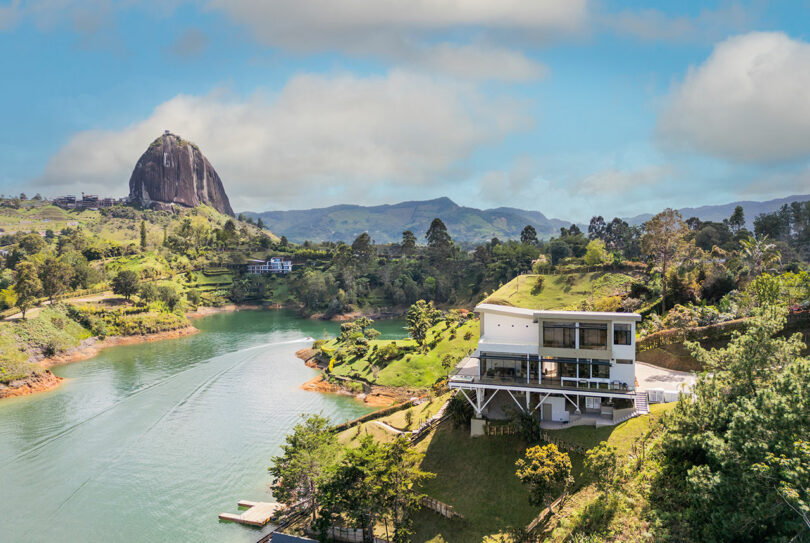 Scenic view of a large rock formation by a river with modern, multi-story buildings and lush greenery under a clear blue sky.