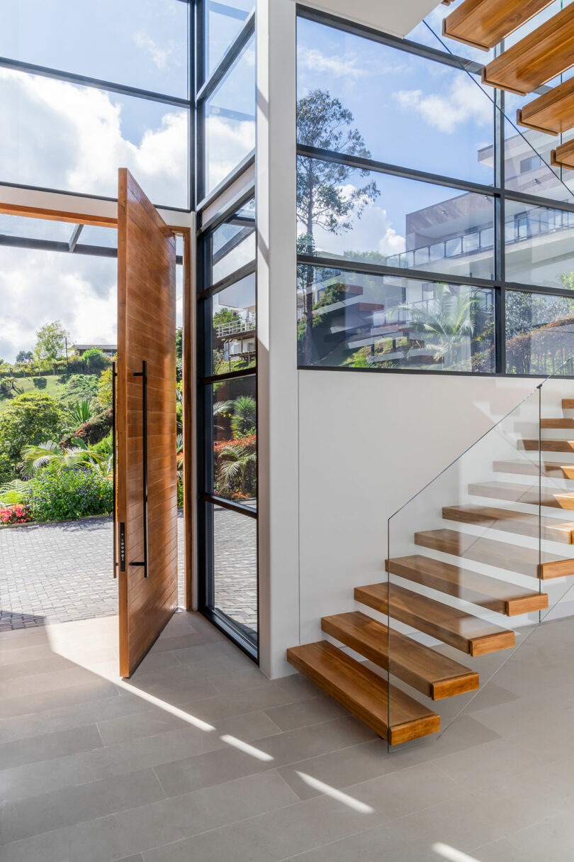 Open door of a modern house with clear glass walls, showcasing an interior floating wooden staircase and a view of lush greenery outside.