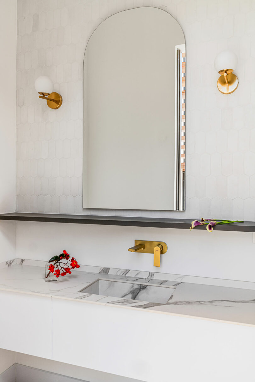 Modern bathroom vanity featuring a marble countertop, arched mirror, gold faucet, and sconce lights, decorated with red and purple flowers.