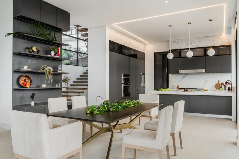 Modern kitchen with a marble backsplash, dark cabinetry, a wooden dining table, chairs, and decorative plants, illuminated by natural light and stylish ceiling lights.