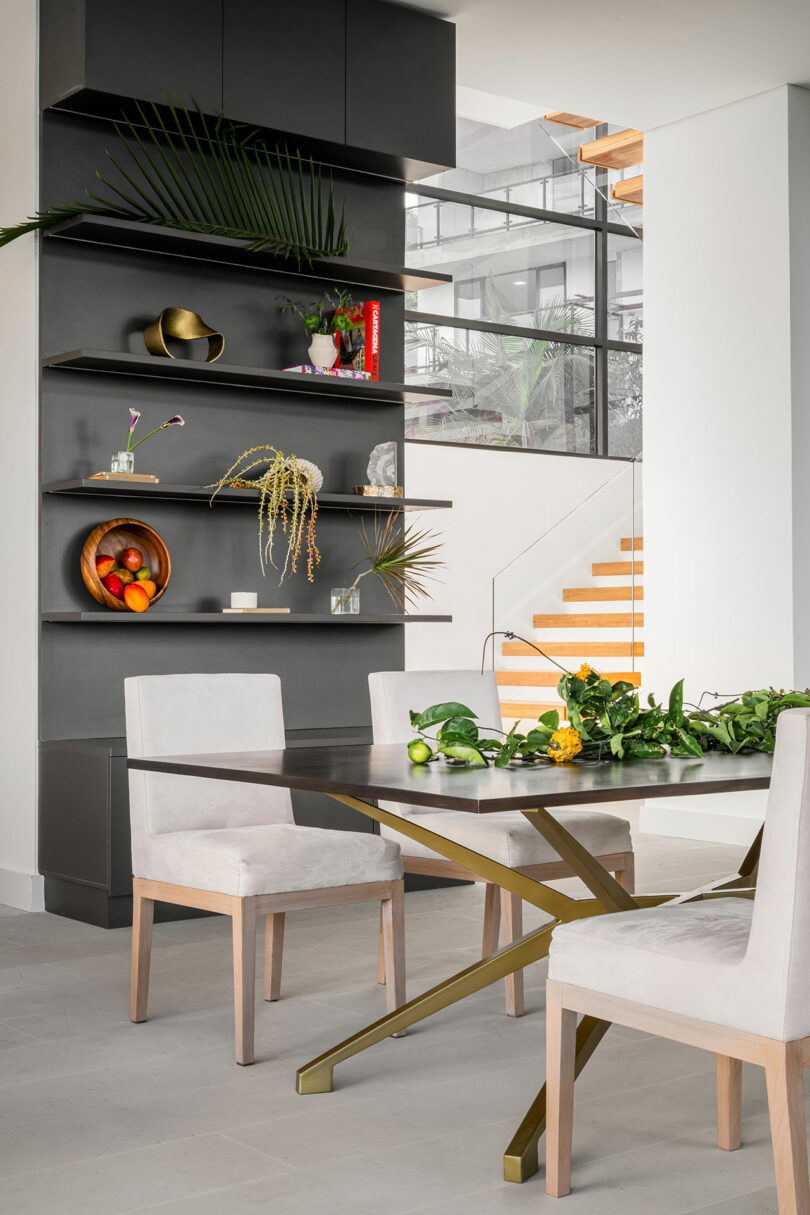 Modern dining room with a black shelving unit, a gold and glass dining table, and four beige chairs, overlooking a staircase.