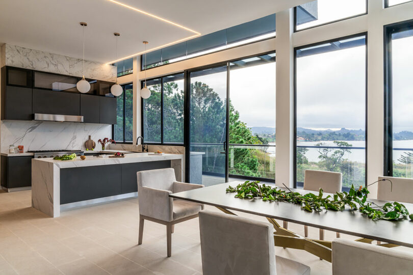 Modern kitchen and dining area with large windows overlooking a scenic view, featuring a sleek design with marble accents and pendant lighting.