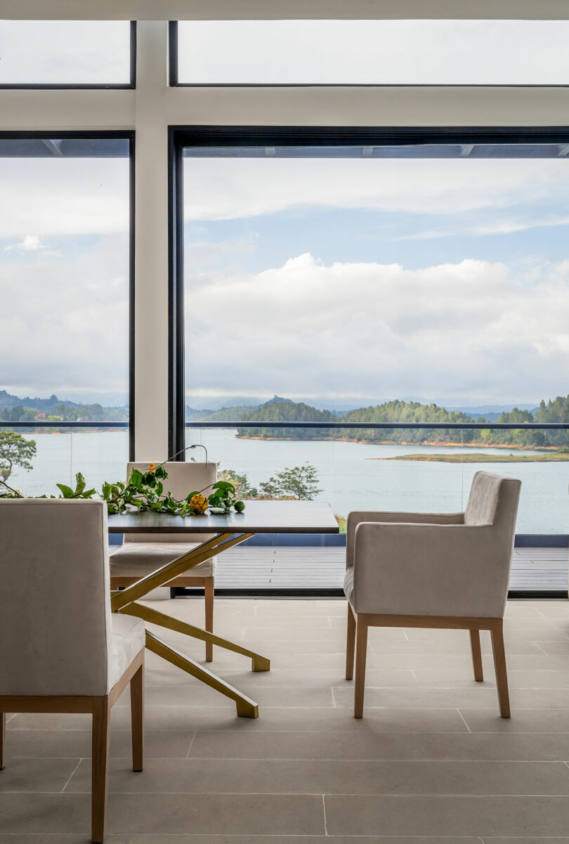 A modern living room with large windows overlooking a scenic lake and hills, featuring a plush chair, a wooden side table, and potted plants.