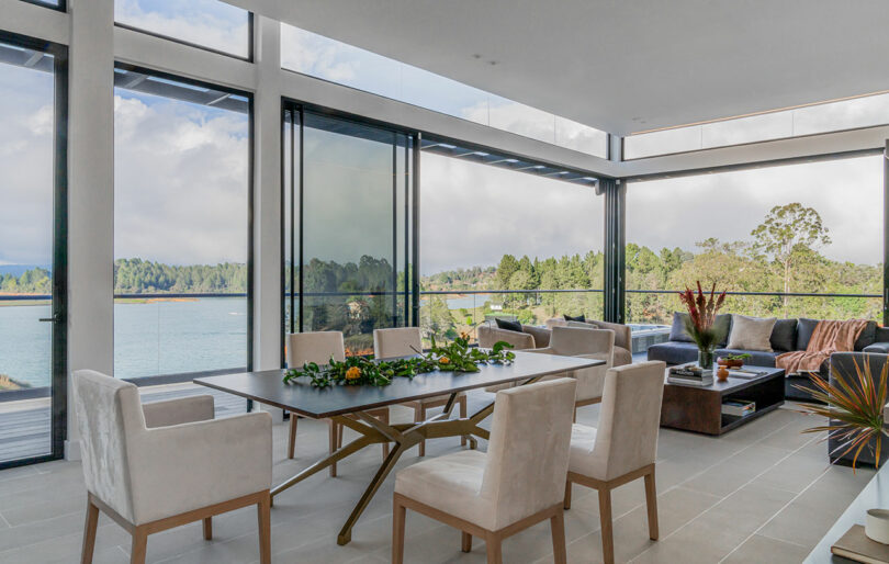 Modern dining room with large windows overlooking a lake, featuring a wooden table, light-colored chairs, and a matching sofa set.