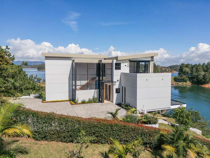 Modern two-story house with large windows and flat roofs, situated by a lake surrounded by a landscaped garden and clear skies.