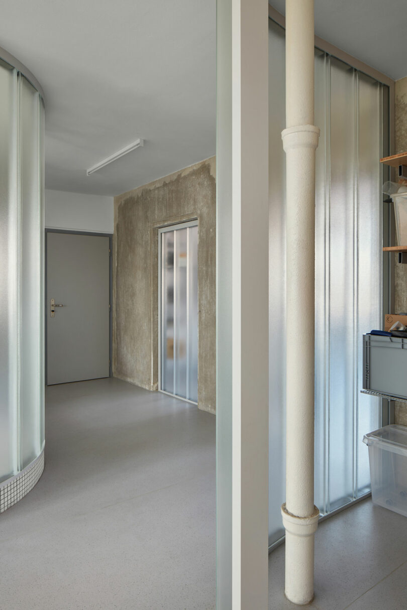 A modern, minimalistic indoor space with frosted glass partitions, a metal door, and a textured concrete wall. There are shelves with storage boxes on the right and a curved wall on the left.