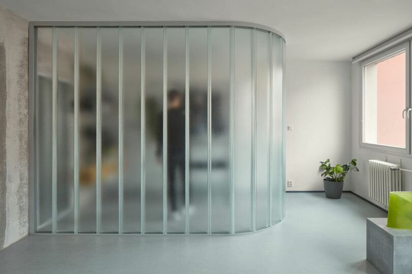 A person blurred behind a frosted glass partition in a minimally furnished room with a large window, a potted plant, a radiator, and a green stool.