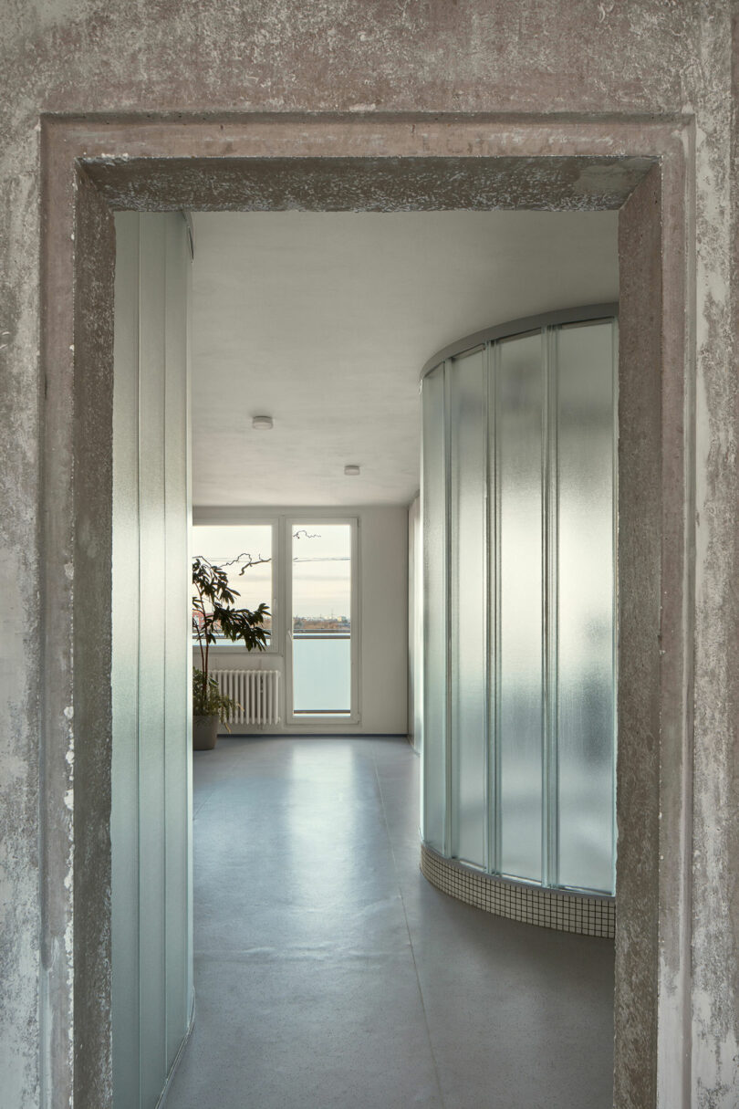 A concrete doorway leads to a minimalistic room with frosted glass partitions, a potted plant, and a large window offering a view of water and distant landscape.