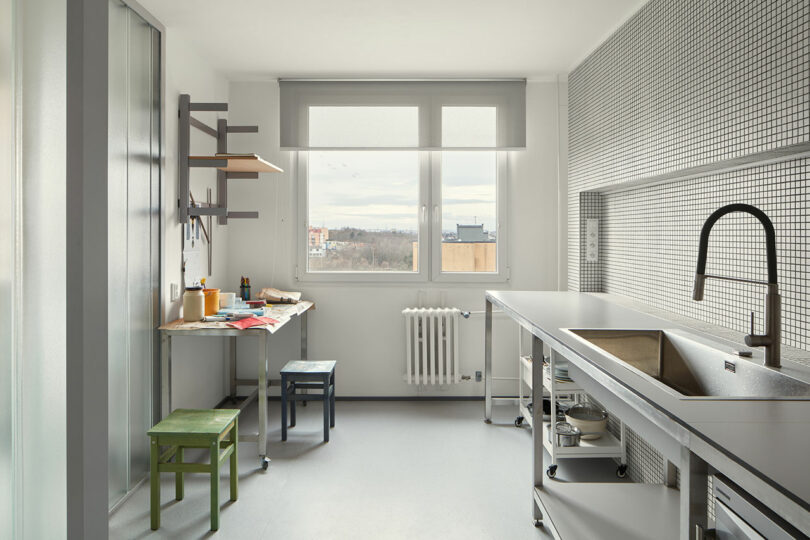 A modern kitchen with a metal sink, tiled backsplash, various stools, a small table with utensils, and a window with a view.