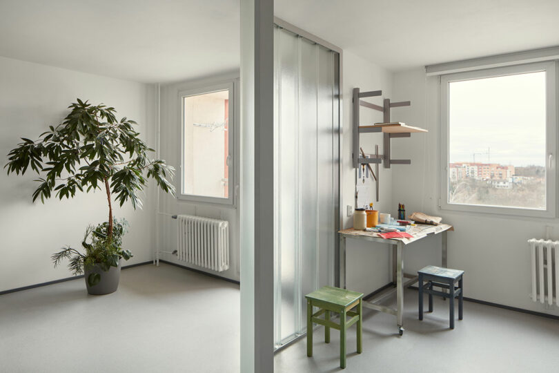 A minimalist room with two windows, a large potted plant, a folding table with art supplies, and two small stools. There is a frosted glass partition and white walls.