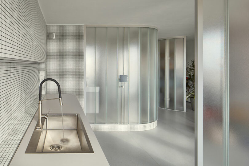 A modern kitchen with a stainless steel sink and faucet, minimalistic design, white tiled walls, and frosted glass partitions.