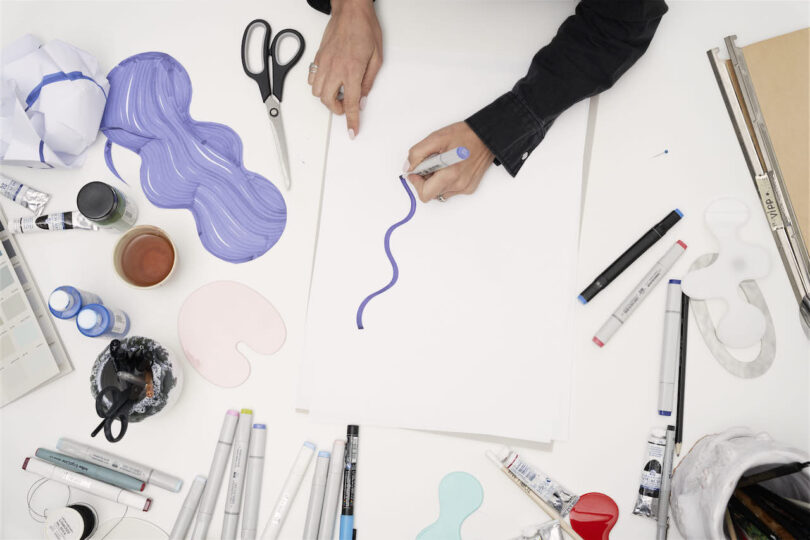 Aerial view of a creative workspace with hands drawing a blue line on paper, surrounded by colorful art supplies and scissors