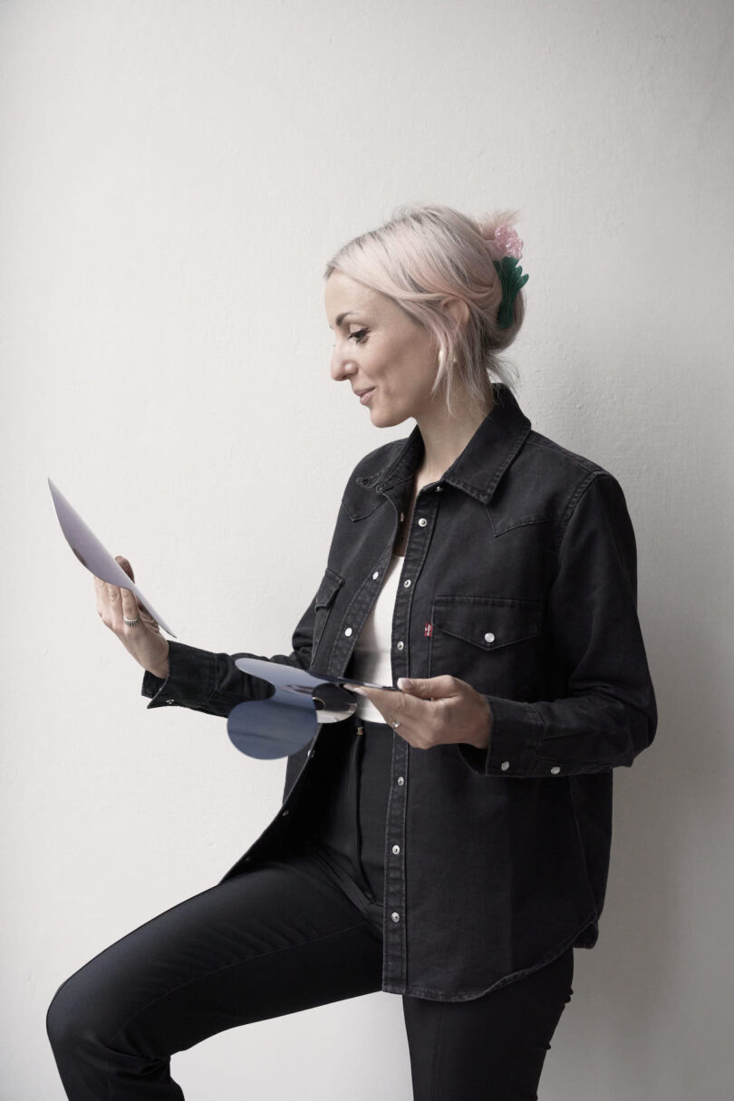 A woman wearing a black shirt, smiles while looking at a magnet in her hands, standing against a plain light background.