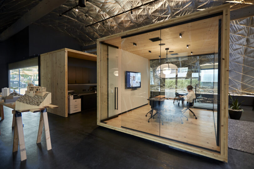 Modern office interior featuring a large wooden cubicle with glass walls, a conference area inside, and architectural models on display