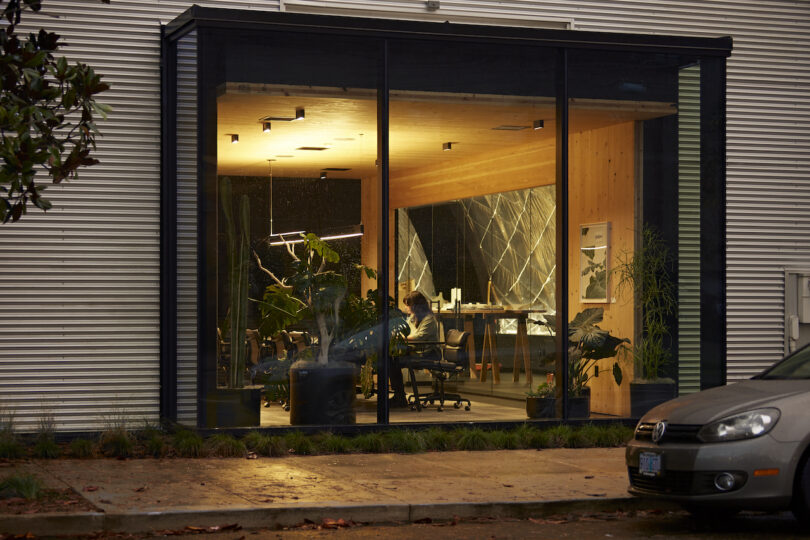 A person working late at night in a modern office with large glass windows, illuminated interior, and visible green plants. a parked car is in the foreground