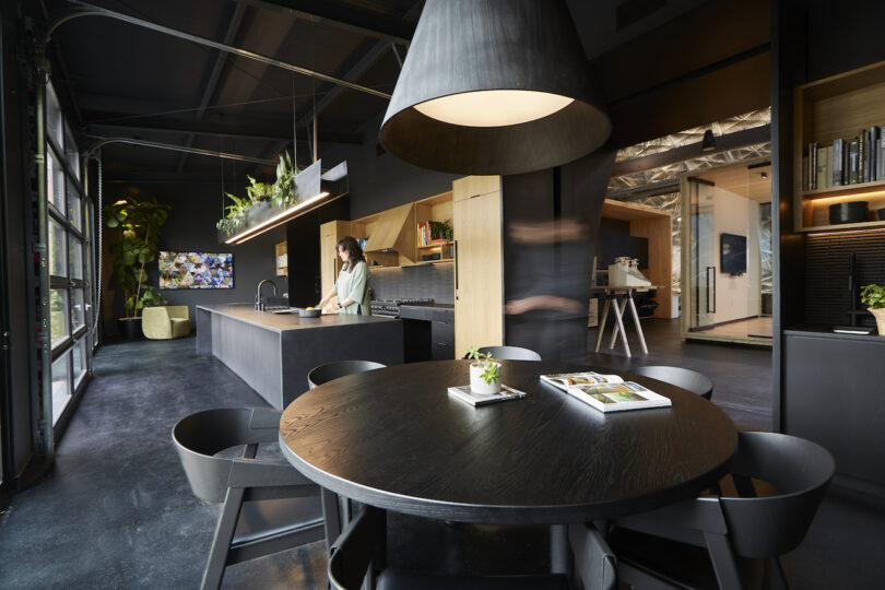 Modern office kitchen with large round table, oversized pendant light, and blurred figure walking by. a woman stands at the counter in the background