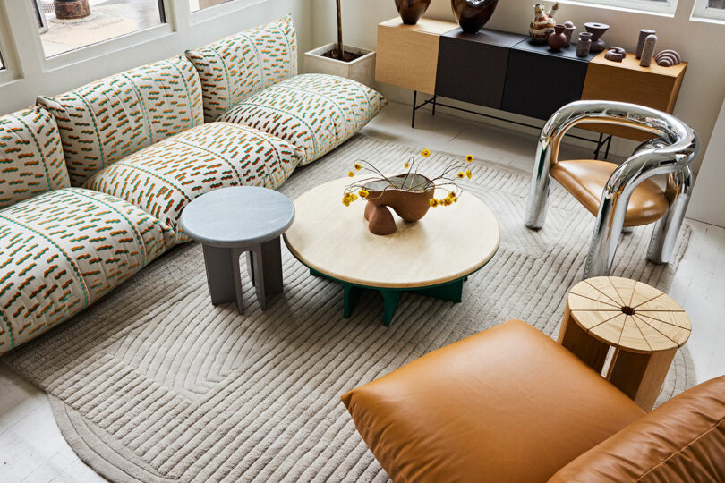 A cozy living space with patterned sofas, two round coffee tables, and a modern chair, accented with decorative vases and a leather pouf, illuminated by natural light.