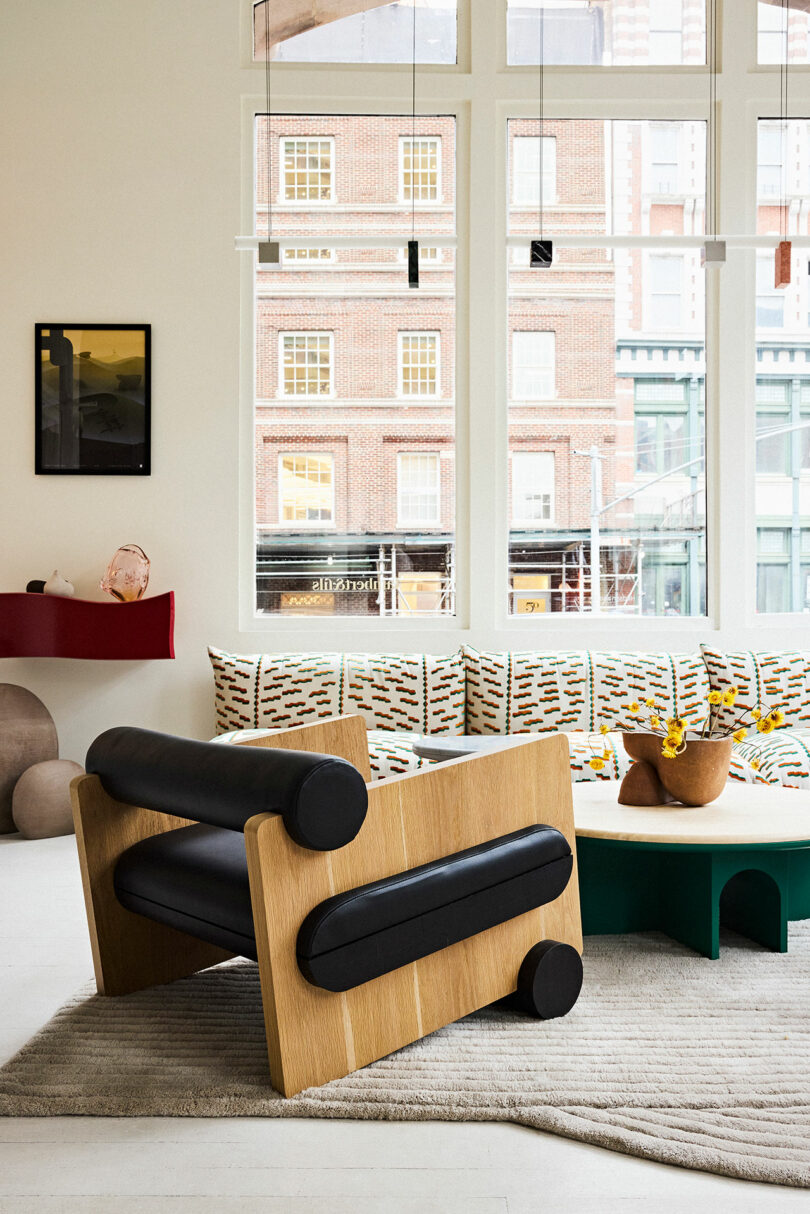 Modern living room with a stylish black and wooden armchair, colorful geometric-patterned sofa, and large windows overlooking city buildings.