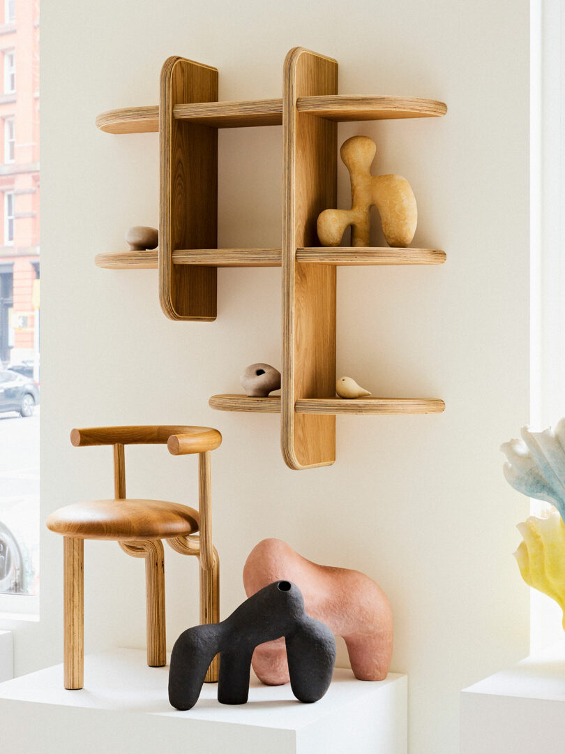 A wooden grid shelf on a wall displaying abstract sculptures, with two artistic chairs and additional sculptures on platforms below.