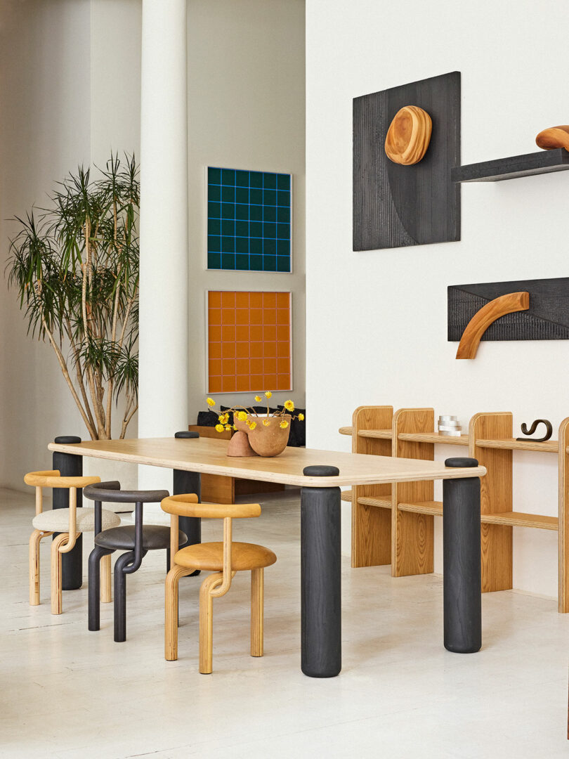 Modern dining room interior featuring a long wooden table with various chairs, colorful geometric wall art, and a tall potted plant.