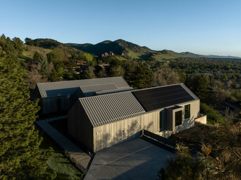 Aerial view of Villa H with a metal roof and solar panels, nestled in a forested area with mountains in the background.