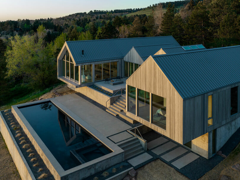 Modern villa with a gabled roof and large windows, surrounded by wooded area, featuring a rectangular pool in the foreground.