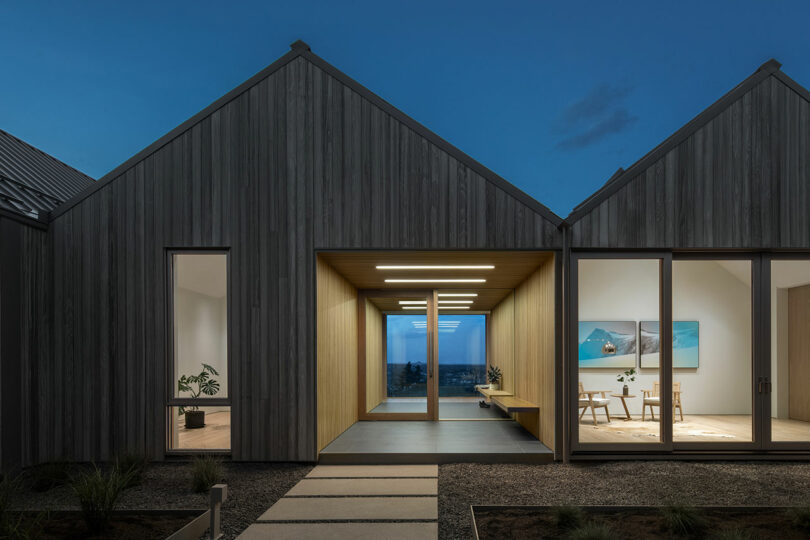 Modern twin-gabled villa at twilight with a central walkway leading to a distant sea view, lit interior rooms visible on either side.